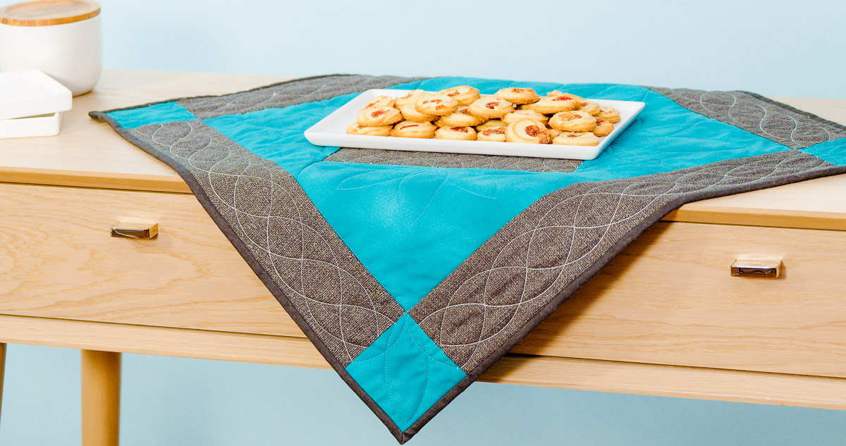 The finished teal and gray Curvy Quilting Table Topper on a wooden table with a tray of cookies arranged beautifully on top.