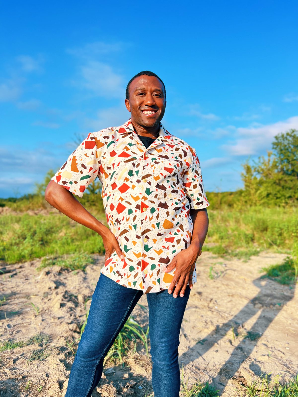 Person standing outdoors in a colorful handmade buttonup shirt with a terrazzo inspired print, paired with blue jeans, smiling under a bright blue sky.