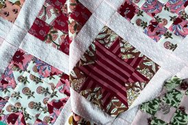 A close up of a quilt in progress. The quilt features block prints arranged in alternating quilt blocks of 16-patches and sawtooth stars. The colors are mostly in shades of pink and red.
