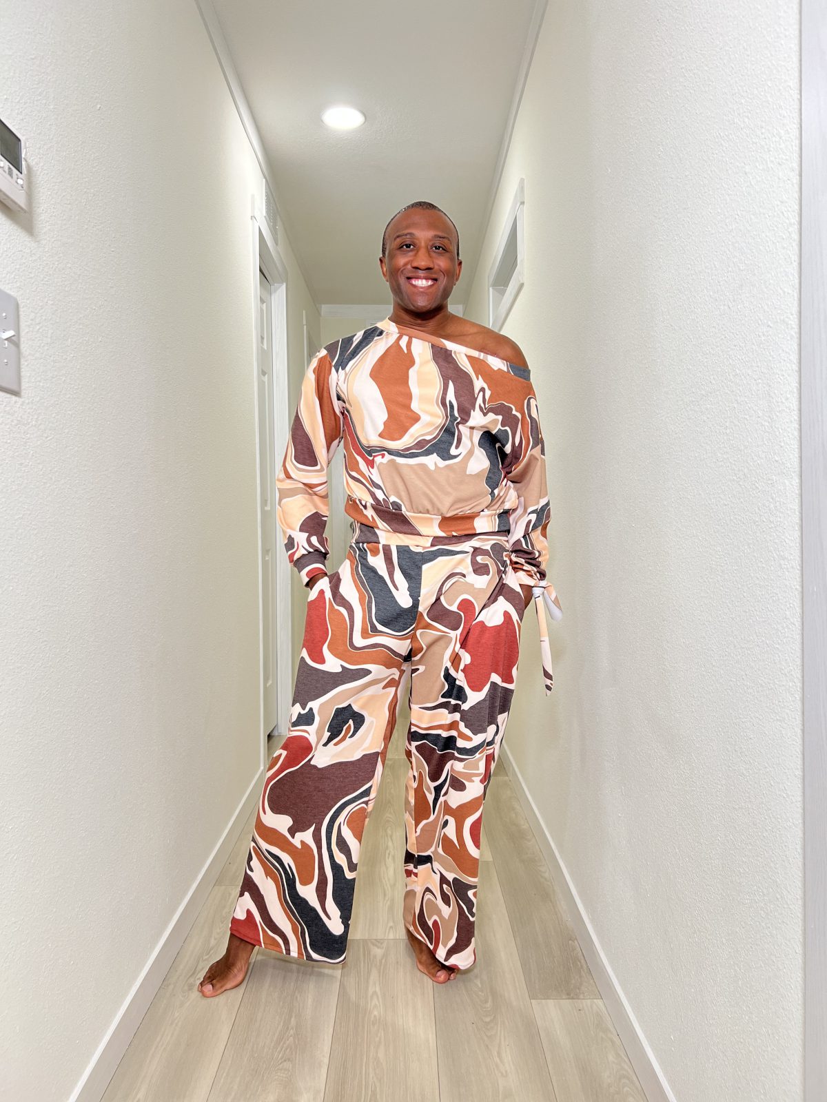 Black man standing in a bright hallway wearing a bold, abstract print sassy sweatsuit with an off the shoulder long sleeve top and matching wide leg pants, smiling confidently.