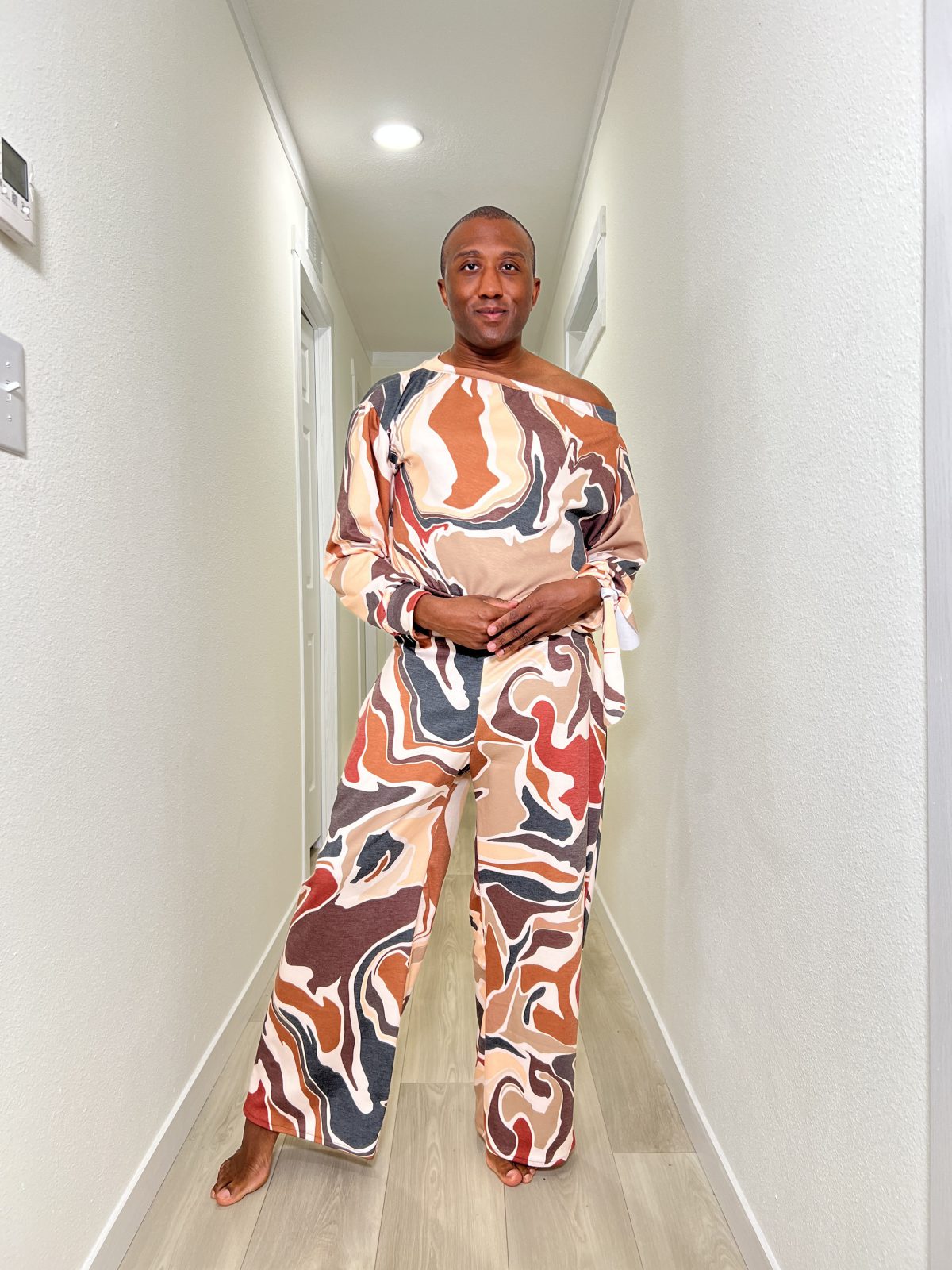 Black man standing in a bright hallway wearing a bold, abstract print sassy sweatsuit with an off the shoulder long sleeve top and matching wide leg pants, smiling confidently.